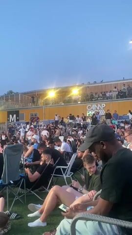 The video shows a crowd of people gathered outdoors, possibly at an event or concert. The sky is a deep blue, indicating it is either dusk or twilight. In the background, there is a yellow building with multiple levels, and lights are visible on the building and on poles. The crowd is seated and standing, with some people in the foreground looking at their phones. The overall impression is of a social gathering in an open-air venue.