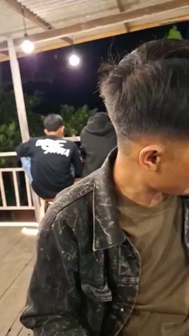 The video opens with a close-up of a young person's head and shoulders, their hair styled in a modern cut. The person is wearing a dark denim jacket over a green t-shirt. The camera then pans to the left, revealing a glass of amber liquid on a concrete ledge, with a plate of food to its left. The background shows a dimly lit outdoor seating area at night, illuminated by string lights. The scene then widens to show a multi-level structure, possibly a restaurant or cafe, with people seated at tables and walking around. The lower level has several small thatched-roof huts and outdoor seating with white tables and benches. The upper levels are more brightly lit with decorative lights. A staircase leads up to the second floor. Several motorcycles are parked in the background. The overall atmosphere is casual and social, with people enjoying their evening.