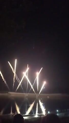 This is a video of a fireworks display over a body of water at night. The fireworks are white and golden, bursting in the sky and reflecting on the water's surface. Several lights are visible in the distance along the shoreline, possibly from buildings or streetlights. Silhouettes of people can be seen in the foreground, indicating they are watching the display. The sky is dark, suggesting it is nighttime.