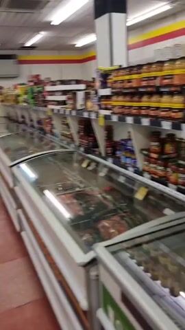 This media file captures a panning shot of the interior of a supermarket in Stonehaven, United Kingdom. The aisles are stocked with a variety of food products, including canned goods, jars of sauces, packaged meals, and fresh produce like onions and what appear to be greens. There are also chilled and frozen display cabinets visible, showcasing more packaged food items.

A man wearing a black hoodie, black tracksuit bottoms, a white baseball cap, and sneakers is the primary human subject. He is seen from behind, pushing a shopping trolley filled with groceries. His presence suggests he is actively shopping.

The supermarket appears well-lit, with fluorescent lighting overhead. The shelves are organized and filled, indicating a typical shopping environment. There are price tags and promotional signs visible throughout, with some prices clearly readable, such as "£1.99". Some text is visible on product packaging, including what looks like "STEAM MEALS" on a display freezer.

The overall atmosphere suggests a normal daytime shopping trip. There are no obvious indicators of the weather outside or the specific time of day beyond it being during operating hours. The scene is devoid of any strong emotional cues.