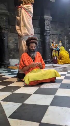 A bearded man with long, dark hair styled in a bun, wearing an orange robe and sitting cross-legged on a yellow cloth, is shown threading beads. He is in a room with a checkered floor and stone walls. Two pillars are visible, one draped with a white cloth and red trim. In the background, a person in a yellow garment is kneeling before an altar with religious figures. The man is focused on his task, his hands busy with the beads and string. The atmosphere appears to be that of a temple or sacred space.