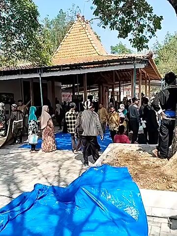 The video shows an outdoor gathering of people in front of a building with a distinctive triangular roof. The building appears to be a traditional structure with open sides supported by pillars. A large blue tarp is spread on the ground in the foreground, and another blue tarp is visible in the background. A group of people are standing around and interacting, some are wearing traditional clothing. There are trees and foliage in the background, suggesting a rural or park-like setting. There is a tall, conical object wrapped in white material on a red box in the middle ground. The overall impression is of a community event or gathering.