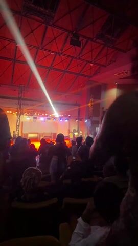 This video captures an energetic indoor stage performance or concert. Several male performers are on stage, illuminated by vibrant, dynamic lighting featuring beams of orange, white, blue, and green. One central figure in a white sleeveless top stands before a DJ setup, actively gesturing to the audience. To his left, another man in a yellow long-sleeved shirt stands, while to his right, a man in a white shirt and black cap is positioned behind a laptop, holding drumsticks. Large speakers, stage lighting trusses, and various electronic equipment are visible on the stage.

A crowd of people, mostly silhouetted from behind, fills the foreground, observing the performance. The setting appears to be a large hall, with faint white text visible on a screen or wall in the background, which includes "OLOWO U..." and "RSITY," likely indicating "Obafemi Awolowo University," aligning with the location context of Ife, Nigeria. A smaller sign on the right also shows "HALL." The event is taking place indoors, likely at night, given the reliance on artificial stage lighting. The overall atmosphere suggests an engaging musical or entertainment event.