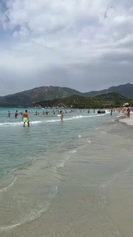 A wide shot captures a crowded beach scene on a cloudy day. The foreground shows the sandy shore with gentle waves lapping at the beach. In the middle ground, the turquoise water is populated with numerous people swimming, wading, and playing. Some individuals are closer to the shore, while others are further out. The background features a line of green, hilly mountains under a grey, overcast sky. The overall impression is a lively beach with many beachgoers enjoying the water despite the cloudy weather.