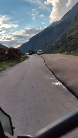 This video shows a road winding through a mountainous region. The road appears to be asphalt, with some water or wet patches on it. On the side of the road, there is a rocky slope covered in some vegetation. In the distance, there are two vehicles on the road. One is a large orange construction vehicle, possibly a bulldozer or front-end loader. The other is a white SUV. There are also a few people visible near the vehicles. The sky above is blue with white, fluffy clouds, and the sun is shining, casting shadows. The overall impression is of a remote, scenic location with ongoing road work or maintenance.