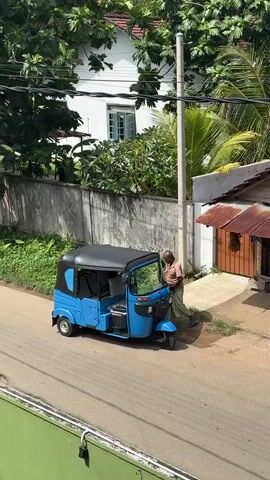 The video shows a blue three-wheeled auto-rickshaw parked on a paved road. A person is partially visible inside the auto-rickshaw, in the driver's seat, and appears to be leaning forward. The auto-rickshaw is positioned at an angle, facing towards the right side of the frame. The background includes a concrete wall, lush green vegetation, a white building with windows, and a utility pole. The scene is illuminated by natural daylight, suggesting an outdoor setting. The camera angle is from an elevated position, looking down at the auto-rickshaw and its surroundings.