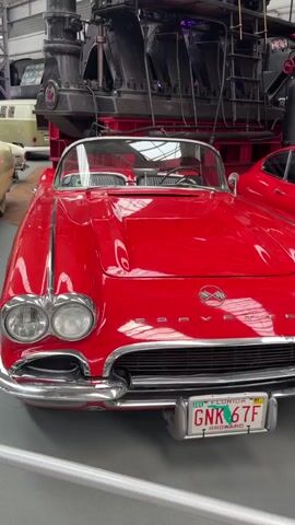 The video pans across the front of several classic cars displayed indoors. The primary focus is on a bright red convertible, likely a Corvette, with its chrome grille and headlights prominently featured. To its left, a cream-colored classic Cadillac is visible, also showcasing its chrome details and large grille. In the background, a portion of a large, dark industrial machine, possibly a train engine, is visible, along with parts of other vehicles and the interior of a building with a high ceiling and industrial lighting. The camera moves smoothly, providing different angles of the cars and their surroundings.