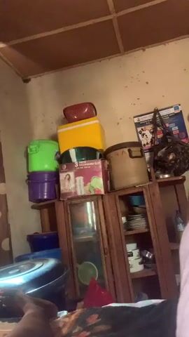 The video shows a bedroom from a first-person perspective. The room appears cluttered, with various items stacked on shelves and on the floor. There are several storage containers, including plastic bins and a cooler. A dresser or cabinet with glass doors holds plates and other kitchenware. Posters and papers are attached to the walls. A window with patterned curtains is visible, and sunlight streams through. The camera pans across the room, briefly showing a person's leg and foot. The overall impression is of a lived-in, somewhat disorganized space.