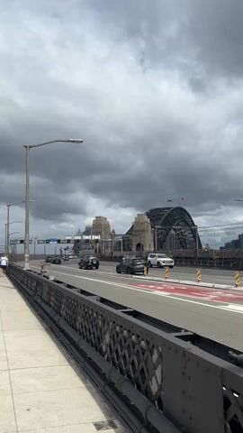 The camera is oriented facing towards the Sydney Harbour Bridge and traffic traveling in the same direction. On the left side of the frame is a sidewalk section with a black metal railing, separating the pedestrian walkway from the car lanes. A person wearing a white shirt and shorts is walking on the sidewalk to the left of the frame. Another person in a white shirt, a grey hat, and a black sweater tied around the neck is seen holding a small brown paper bag and standing on the sidewalk on the left side of the frame. The Sydney Harbour Bridge is shown in the background, with several cars traveling on it. Dark storm clouds are overhead.