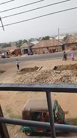 The scene unfolds from an elevated perspective, possibly a balcony, overlooking a modest townscape. A paved road cuts through the scene, with sparse vehicular traffic including a motorcycle and a three-wheeled vehicle parked below the camera. A few individuals stroll along the road and its shoulder. One person is clad in a pink garment. A brown goat saunters across the unpaved ground in the foreground. In the background, various buildings with low-slung roofs, trees, and a distant, hazy horizon form the backdrop. An overhead network of utility wires and the metal railing of the balcony provide a frame for the scene.
