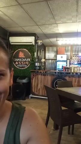 The video shows the interior of a pizza restaurant, likely named "Racing Pizza," based on the logo visible on the wall. The restaurant features rustic decor, including corrugated metal walls behind the counter, wooden accents, and a flat-screen TV playing what appears to be a baseball game. There are tables and chairs set up for dining. The video begins with a woman smiling in the foreground, partially obscuring the view, but then pans to show more of the restaurant interior. There are no visible interactions or actions, only the static scene of the restaurant.
