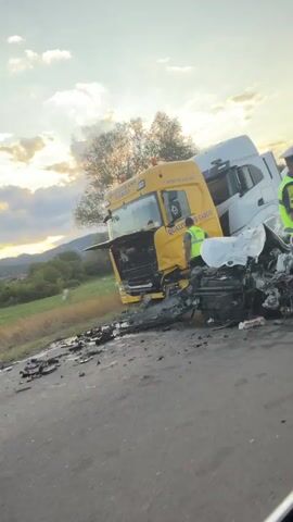 A police officer in uniform with a reflective vest holds up a stop sign to direct traffic at the scene of a multi-vehicle accident. The accident involves at least two semi-trucks and several cars. The front of a yellow semi-truck appears significantly damaged, and it is connected to a white semi-truck. A white car in the foreground is also heavily damaged. Another vehicle, an orange flatbed truck, drives past the accident scene. The accident appears to have occurred on a paved road in a rural area with trees and hills in the background. The sky is cloudy with the sun setting.