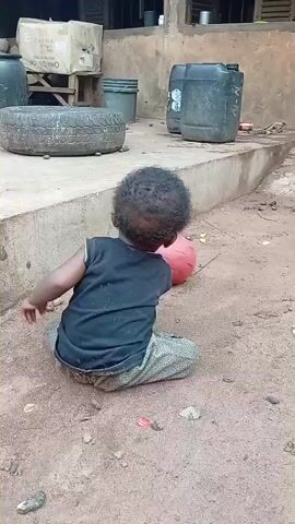 This short video depicts a young child playing with a pink ball in a dusty, outdoor setting. The child, viewed from behind, is wearing a dark t-shirt and patterned trousers and is seated on the ground. They appear to be interacting with the ball, possibly pushing or rolling it with their hands or a stick.

The background reveals a somewhat dilapidated outdoor area, possibly a yard or courtyard. Visible objects include large plastic barrels, a discarded tire, crates or boxes, and a bucket. A building with an open front, likely a house or shelter, is present in the background, with laundry hanging on a line. The ground is predominantly dirt and dust, with some scattered debris.

The lighting suggests it is daytime, though the exact time is unclear. The overall impression is one of a simple, everyday scene in a rural or less developed area. There is no discernible text in the video. The child's actions suggest a moment of solitary play.