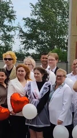 The video shows a group of people posing for a photo. In the front row, there is a girl with a white sash that says "graduate" in Russian. She is wearing a dark dress with a white lace apron and holding a white balloon. Next to her, there are two women in white jackets. One of them is holding a red heart-shaped balloon, and the other is wearing glasses. In the back row, there are several young men with different hairstyles and facial expressions. One of them has blonde hair and sunglasses. There is also a man with dark hair and a white shirt who is smiling at the camera. The background includes trees and a building. The overall impression is that this is a graduation ceremony or celebration.