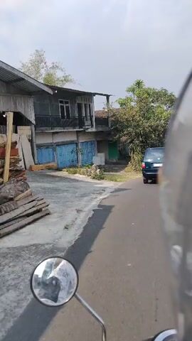 The video shows a first-person perspective of someone riding a motorcycle down a paved road. The road appears to be in a rural or semi-rural area, with houses and vegetation visible on either side. A blue minivan is driving ahead of the motorcycle. The houses are simple in design, some with balconies and tiled roofs. There are trees and palm trees in the background, and the sky is overcast. The rider's hand is visible in the bottom right corner of the frame, holding the handlebars. The reflection in the motorcycle's mirror shows the rider's helmet and part of their face. The scene is lit by daylight.