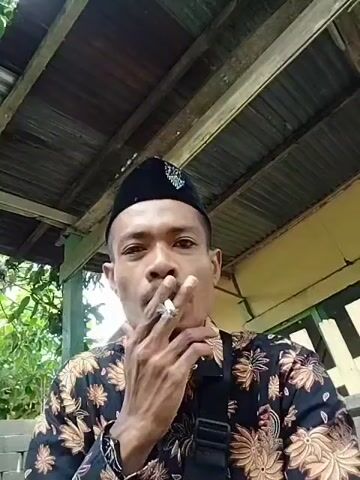 The video features a man from the chest up, looking directly at the camera. He is wearing a black songkok (a traditional cap) and a dark-colored, long-sleeved shirt adorned with light brown floral patterns, characteristic of batik. A black sling bag is visible across his chest.

At the start of the video, the man has a lit cigarette in his mouth, takes a drag, and exhales smoke. He then removes the cigarette from his mouth, holding it in his right hand, and maintains a neutral expression while continuing to face the camera.

The setting appears to be an outdoor or semi-open area. Above him, a corrugated metal roof supported by wooden beams is visible. The background shows green foliage and a light-colored building, possibly yellow, with white vertical details. The lighting suggests it is daytime. There is no visible text.