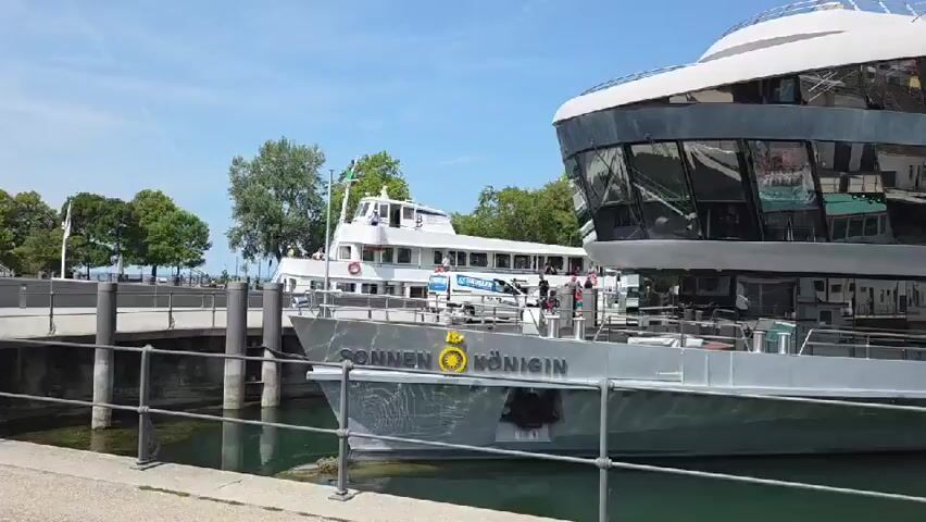 This is a daylight video of a boat docked at a harbor. The boat in the foreground has the name SONNEN KÖNIGIN on its side. The boat behind it has a white upper deck with people visible. Another smaller boat is docked to the right of the SONNEN KÖNIGIN. The water is calm and a blue sky with some clouds is visible. Trees and buildings are visible in the background.