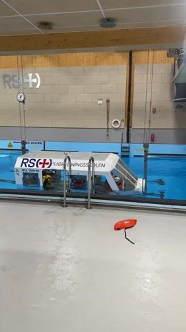 An indoor swimming pool, likely a maritime training facility in Horten, Norway, is the setting for a simulated emergency exercise. A large, white, boat-like structure, labeled "RS(+) SJØREDNINGSSKOLEN" and "WLL 1000 KG", is performing a controlled capsize or submersion. The structure slowly tilts and sinks into the blue pool water, with several individuals visible inside or near it as it submerges. The pool walls are tiled with wooden paneling above, and feature signs including "NO DIVING" and "5,0m" depth markers. A red buoy or life preserver lies on the wet concrete floor in the foreground, next to the pool. A clock is visible on the back wall.