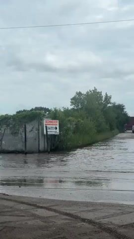 A white semi-truck with a red shipping container is driving through a flooded road. The water is approximately up to the truck's axles. The road appears to be paved with tire tracks visible on the dry portions. Trees and a fence line the sides of the road. The sky is overcast. Another vehicle is visible in the distance, also in the flooded area.