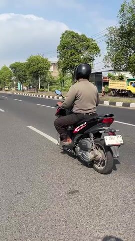 This is a vertical video clip of a person riding a motorcycle on a road in Siring, Indonesia. The rider is wearing a black helmet and a grey jacket. The road is paved and has white lane markings. There are trees and buildings on the side of the road, and the sky is partly cloudy. The sun is shining, suggesting it is daytime. The video appears to be filmed from another motorcycle, showing the road ahead and a portion of the handlebars in the foreground. Another motorcycle is visible in the distance. The overall mood is casual and the activity is transportation.