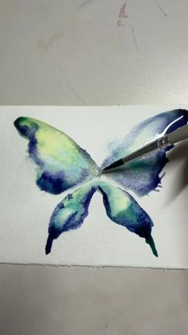 A person's hand holding a paintbrush is adding red watercolor to the center of a painted butterfly. The butterfly's wings are painted with shades of blue, green, and yellow watercolor in a loose, blended style. The painting is on white paper, which is resting on a light-colored surface.