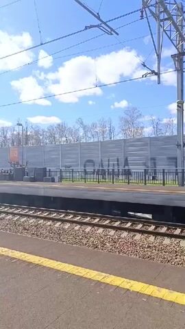 The video shows a view of a train platform on a sunny day. The camera is positioned at ground level, looking across the train tracks towards the platform. There is a yellow safety line on the edge of the platform. The train tracks are filled with gravel. A low metal fence runs along the edge of the platform. Behind the fence, there is a tall, corrugated metal wall that is presumably a sound barrier. A sign with white lettering indicates that the platform is for the "Skolkovo" stop. The sky is blue with scattered white clouds. Overhead, there are electrical wires and supporting metal structures. The background includes some bare trees. The overall impression is of a quiet, rural train station.