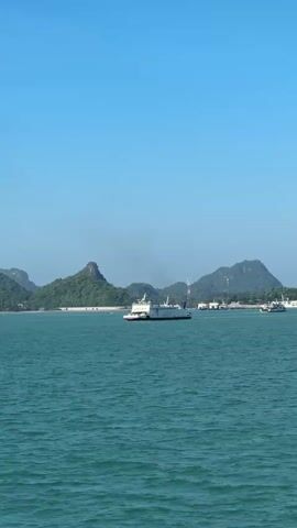 The video shows a wide shot of a body of water, likely the ocean, under a clear blue sky. In the distance, there are several green, hilly islands or land masses. A ferry boat is visible in the middle of the water, traveling from left to right. Another, smaller boat is further to the right. The water is a turquoise green color and appears to be slightly choppy. The overall scene is serene and depicts a calm day at sea.