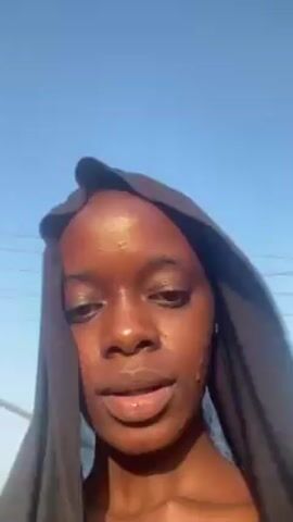 A person with dark skin and brown eyes is filming a selfie video. They are wearing a dark, loose-fitting hood over their head. They are standing outdoors on a sunny day with a clear blue sky. The person is talking to the camera, and their mouth is moving. There are power lines and other indistinct objects in the background. The person's head moves slightly from side to side as they speak.