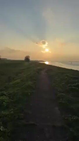 The camera looks down a dark, gravel path at sunset. The path is lined on either side by bright green, grassy embankments. To the right of the path is an expansive, shimmering body of water, reflecting the orange glow of the setting sun. The sky is a mix of light blue and soft orange, with thin clouds partially obscuring the sun. A single, bare tree stands on the left side of the path, further in the distance. The scene is tranquil and serene.