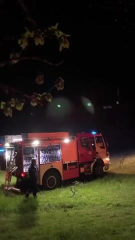 The video captures a nighttime scene in Dolno Drianovo, Bulgaria, where firefighters are actively battling a large fire. A red and white fire truck, illuminated by its flashing blue lights, is parked on a grassy area to the left. Several firefighters are visible; some are in uniform and working near the fire, while others are seen in silhouette, observing the intense orange flames and plumes of smoke emanating from the blaze. The fire appears significant, casting a bright glow across the dark surroundings. Other individuals are also present, standing in the foreground, watching the firefighting efforts. The scene is dark, with light primarily provided by the fire and the vehicle's equipment.
