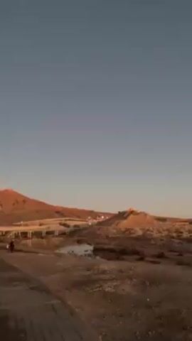 The video depicts a desert landscape under a clear, blue-to-orange gradient sky suggesting early morning or late evening light. A small body of water reflects the sky and nearby structures. A pedestrian walkway is seen in the foreground. A road or bridge cuts across the landscape in the middle distance. The ground is predominantly dry, with sparse vegetation. In the background, there are small buildings or structures on a mound, possibly a village or settlement. A vehicle passes by in the distance.