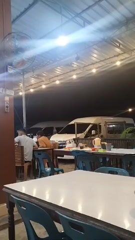 The video shows an open-air dining area at night, likely a casual restaurant or eatery in Bayeun, Indonesia. Several people are seated at tables, some facing away from the camera, while others are partially visible. The tables have white or dark wooden tops, and the chairs are a mix of blue plastic and brown wood.

The area is brightly illuminated by overhead lights and string lights running along the ceiling structure. Ceiling fans are mounted on pillars, suggesting a warm climate. In the background, several white vans are parked, possibly belonging to customers. A prominent brown pillar stands in the foreground on the left, near which a ceiling fan is also attached. The atmosphere appears casual and relaxed, with patrons enjoying their meals or conversations. There is no clearly discernible text visible in the video.