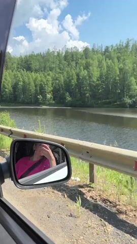 A video captures a view from inside a car, looking out the passenger window. A side-view mirror reflects a young woman wearing a pink shirt and a seatbelt. She is covering her mouth with her hand. Outside the car, a body of water, possibly a lake or river, is visible. The water is surrounded by a dense forest of green trees. The sky is blue with scattered white clouds. A metal guardrail runs along the side of the road, separating it from the embankment. The road itself appears to be unpaved or dirt. The overall scene suggests a scenic drive through a natural, wooded area on a sunny day.