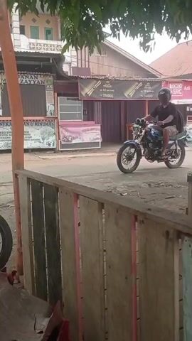A person wearing a black helmet and a black t-shirt rides a blue motorcycle. The person is wearing a black backpack. The motorcycle has a black seat and silver wheels. The person is riding down a dirt road next to a sidewalk. There are trees and buildings on either side of the road. A red motorcycle is parked on the left side of the image. A person wearing a red beanie is looking towards the blue motorcycle. There is a wooden fence in the foreground.