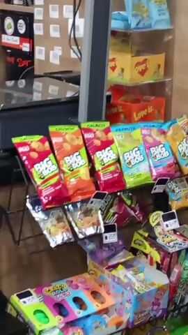 The video shows a variety of snack foods and beverages in a store setting. The first shot shows a display of colorful chip bags near a weighing scale with a smiley face sticker. The camera then pans to a display of different types of dried snacks in clear plastic containers. Finally, the camera shows a refrigerator stocked with various cans of beverages.