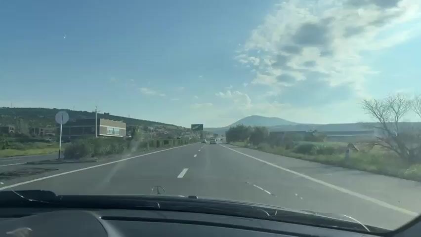 The video shows a first-person view from a moving car on a highway. The car is traveling on the right side of the road. The sky is blue with scattered white clouds. To the left of the highway, there are some buildings, trees, and a hillside with more buildings. There is also a billboard visible on the left. To the right of the highway, there are trees, green vegetation, and fields. In the distance, a mountain is visible under the clouds. Several other vehicles are seen on the highway ahead. The interior of the car's dashboard is partially visible in the foreground. The time stamp indicates the video starts at 00:00 and ends at 00:04.