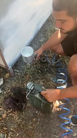 A person is spray painting a white surface. The person is crouching on the ground, holding a spray gun connected to an air compressor hose. The surface being painted appears to be a large, flat panel, possibly metal, with a slight sheen. There is some debris on the ground, including dried leaves and grass. Other objects visible include a green toolbox, a blue coiled hose, and a piece of dark fabric. The lighting suggests it is daytime, possibly outdoors. The person's action is focused on applying paint to the surface.