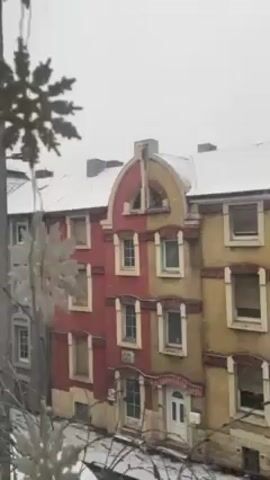 The video shows a winter scene of three buildings on a snowy street. The building on the left is grey, the middle one is red and yellow, and the building on the right is yellow. The roofs are covered in snow. A man is walking on the sidewalk, also covered in snow. Snowflakes are falling. A white decorative snowflake hangs in the foreground, slightly out of focus.