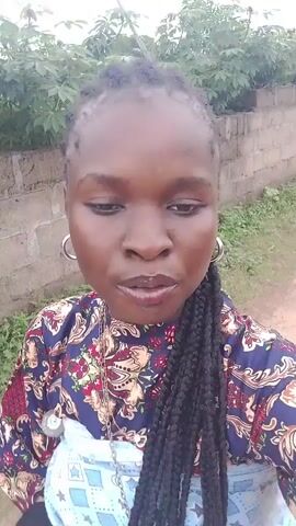 A woman with dark skin and braids is recording a video of herself outdoors. She is wearing a colorful patterned shirt and large hoop earrings. The background appears to be a dirt path next to a low brick wall and some vegetation. She smiles and makes facial expressions throughout the short clip.