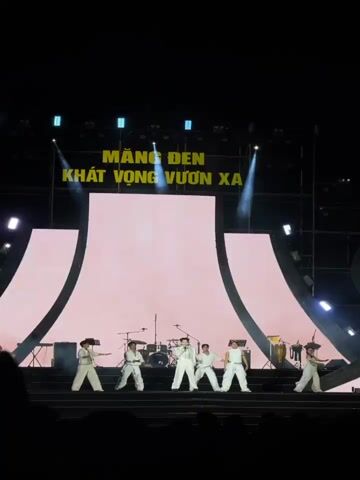 A nighttime concert or performance is taking place on a large, brightly lit stage. Seven performers, all dressed in matching white outfits, are dancing in a synchronized manner across the front of the stage. Behind them, a band with a drum set and other instruments is visible, suggesting live music accompaniment.

The stage features a prominent, tall central screen displaying a vibrant pink and red backdrop with a traditional East Asian aesthetic, including stylized clouds, pagodas, and a large central symbol resembling a longevity or happiness character. Two curved, illuminated structures frame the central screen, adding depth to the set design. Above the stage, a large banner with bold yellow text reads "MĂNG ĐEN KHÁT VỌNG VƯƠN XA," which translates to "Măng Đen - Aspiration to Reach Far," indicating the event's location and theme, likely Măng Đen, Vietnam. Bright spotlights, casting blue and white beams, crisscross the stage from above, while purple lights illuminate the stage edges and structures. The audience area in the foreground is dark, highlighting the energetic performance under the bright stage lights.