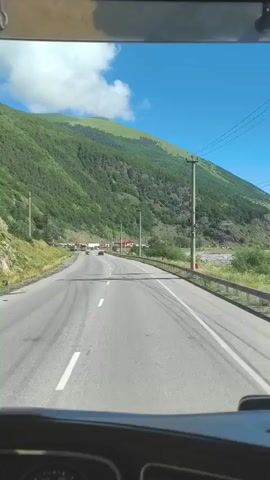The video shows a view from inside a vehicle driving on a paved road. The road is an asphalt two-lane highway with white lane markings. On the left side of the road, there is a grassy embankment leading up to a steep, green, tree-covered mountain. Several utility poles with power lines run along the side of the road. On the right side of the road, there is a metal guardrail and a rocky, shallow riverbed. In the distance, there are a few buildings visible, as well as other vehicles on the road, including cars and trucks. The sky is blue with some scattered white clouds. The weather appears to be clear and sunny. The vehicle's dashboard is partially visible at the bottom of the frame.