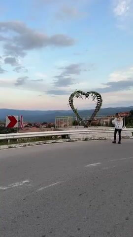 The video shows a young woman with blonde hair, wearing a white hoodie and dark pants, walking near a large heart-shaped sculpture covered in greenery and flowers. The sculpture is situated on a platform overlooking a town nestled in a valley with rolling hills and mountains in the background. The sky is a pale blue with scattered clouds, suggesting it's either early morning or late evening. The woman is captured from behind as she walks towards the sculpture and then stands near it, looking out at the view. A road with white lines is in the foreground. A red and white sign is visible on the left side of the road. The overall scene is peaceful and picturesque.