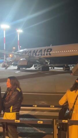 At night, a crowd of people are lined up behind metal barricades in an airport. The lighting is bright, with spotlights on poles that are striped red and white. An airplane is visible in the background. Most of the people are wearing jackets and carrying bags or luggage. They appear to be waiting to enter a building marked “A1.” There is little movement and no discernible interaction between the people.