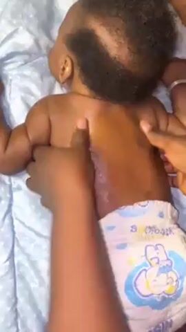 A close-up shot shows a baby lying on its stomach on a light blue blanket. The baby, wearing a diaper with cartoon characters, has dark skin and a small amount of hair. An adult with dark skin is shown massaging the baby's back and neck with oil, using both hands in a gentle, circular motion. The baby remains still during the massage. The focus is on the interaction between the adult and the baby during the massage.