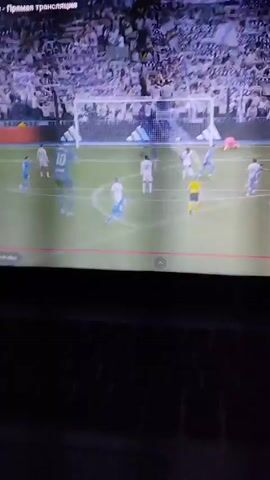 A TV screen shows a replay of a soccer play. Two players jump to head the ball, one wearing white and the other blue. The player in white falls to the ground. The ball continues towards the goal where the goalie is.