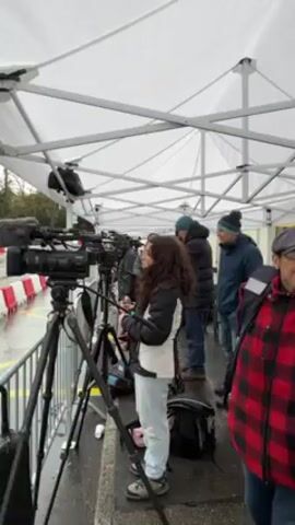 The video shows a group of camera operators and other people standing under a white tent on a rainy day, with several professional video cameras set up on tripods pointing toward a street. The street is lined with red and white barriers and yellow painted lines, indicating a prepared course. On the opposite side of the street is a modern building with a sign saying "INTERCONTINENTAL".  The camera operators appear to be waiting to film something on the street, possibly a race or procession.