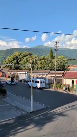 This is a video of a street scene in Şəki, Azerbaijan. The scene is captured from a slightly elevated perspective, looking down onto a road with buildings and trees.

In the foreground, a white van is driving down the road. There are also a few other cars visible, including a white sedan. A traffic light is visible on the left side of the road, showing a green light. A yellow pole with electrical wires runs diagonally across the frame. On the right side of the road, there are low-rise buildings with arched windows, suggesting shops or apartments. Several potted plants are placed in front of these buildings. A few people are visible walking or sitting outside these establishments.

In the middle ground, a dense canopy of green trees dominates the view, with a tall, slender cypress tree standing out amongst them.

In the background, a large, verdant mountain rises up under a bright blue sky with scattered white clouds. The lighting suggests it is daytime, likely late afternoon given the long shadows cast on the road. The overall atmosphere appears peaceful and serene, with the natural landscape providing a beautiful backdrop to the urban setting. There is no visible text in the media file.