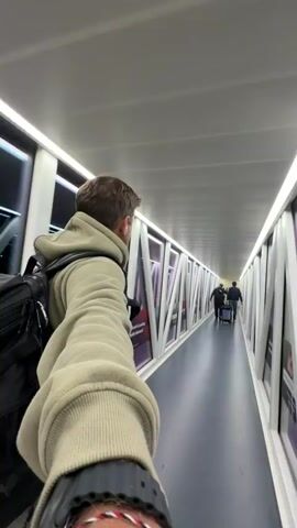 A man is walking down an airport jet bridge, filming himself with his outstretched arm. He is wearing a light beige hoodie, a black backpack, and a large black watch. He is also wearing a white earbud in his right ear. Two other people are walking ahead of him, pulling a suitcase. The jet bridge has white triangular supports along the windows on either side and is well-lit with fluorescent lights. An advertisement for Batelco is visible on the windows of the jet bridge. The floor is a dark blue.