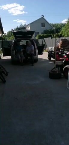 Two men sit in the open trunk of a silver minivan. One man on the left wears a blue t-shirt and shorts. The man on the right wears a light blue striped shirt and dark pants. The minivan has its rear hatch and side doors open. A house and fence are visible in the background. An all-terrain vehicle (ATV) and other objects are parked to the right of the minivan. The scene appears to be outdoors on a paved surface.