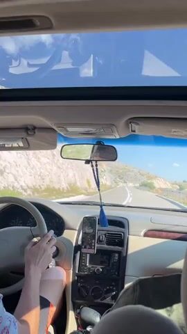 The video shows the interior of a car driving on a road. The perspective is from the passenger seat. A person's arm is visible on the steering wheel. A phone is mounted on the dashboard. A rearview mirror hangs from the windshield. The scenery outside the car shows hills and a blue sky. The video is composed of several short clips, suggesting a continuous journey.